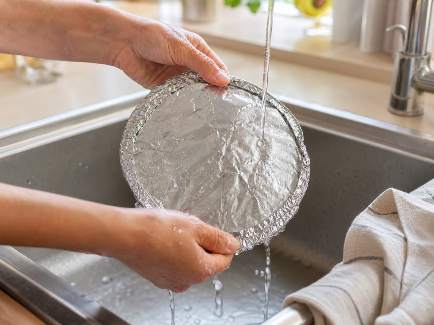 Two hands rinsing a reusable foil food cover in a kitchen sink